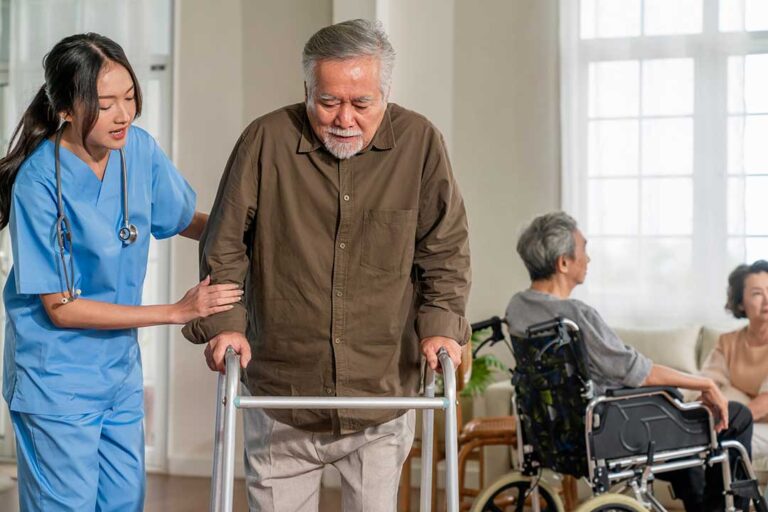 Older adults and staff walk through a bright assisted living hallway with handrails, clear paths, and emergency signage.